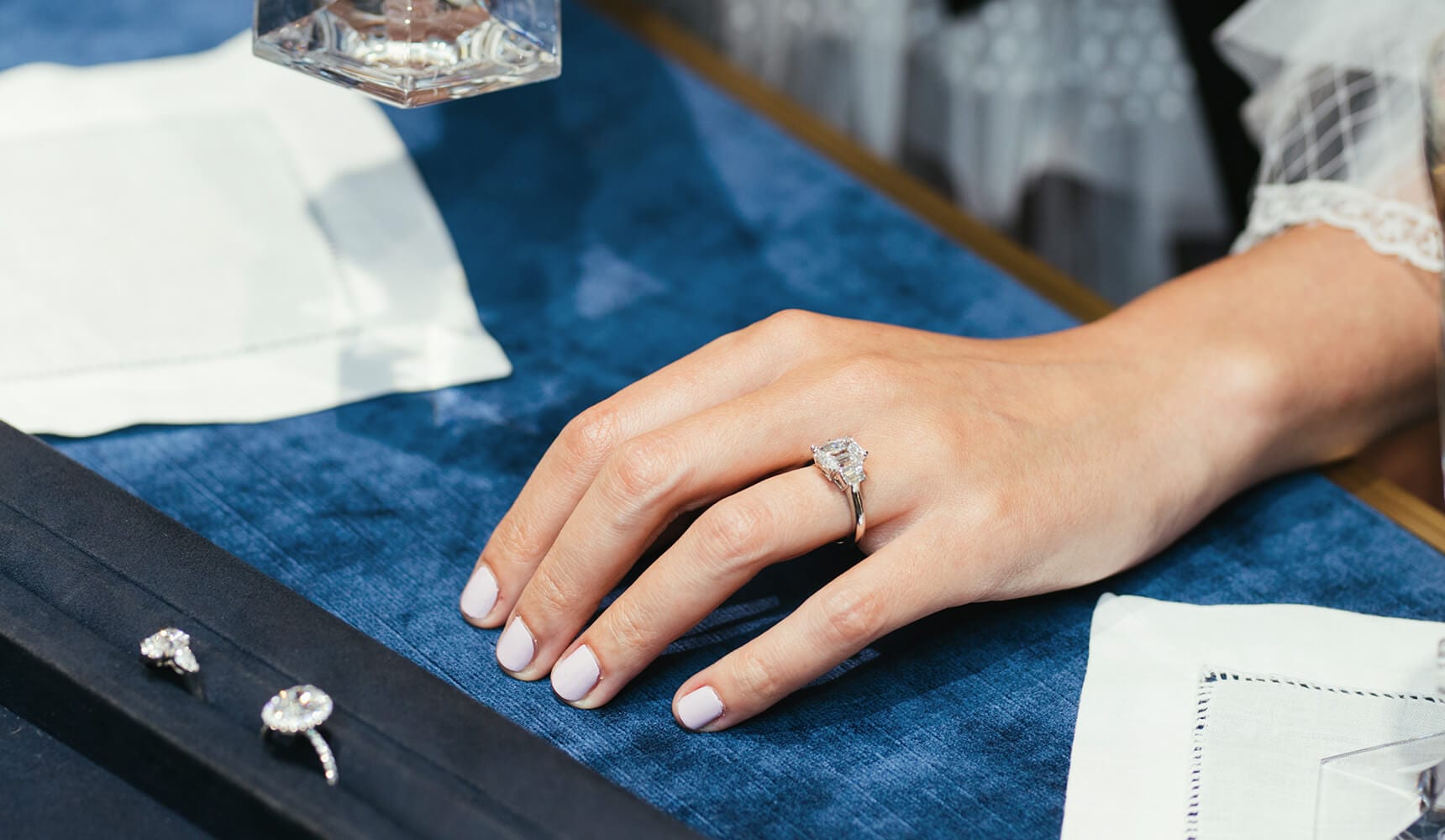 Woman's hand wearing a Harry Winston Classic Winston Emerald-Cut Diamond Engagement Ring next to a tray of Harry Winston Diamond Engagement Rings